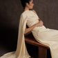 Woman in a white saree sitting on a wooden bench against a dark background
