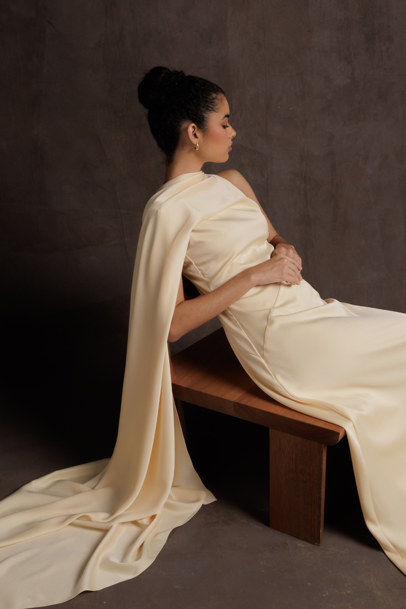 Woman in a white saree sitting on a wooden bench against a dark background