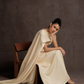 Woman in a flowing beige dress sitting on a wooden chair against a brown background