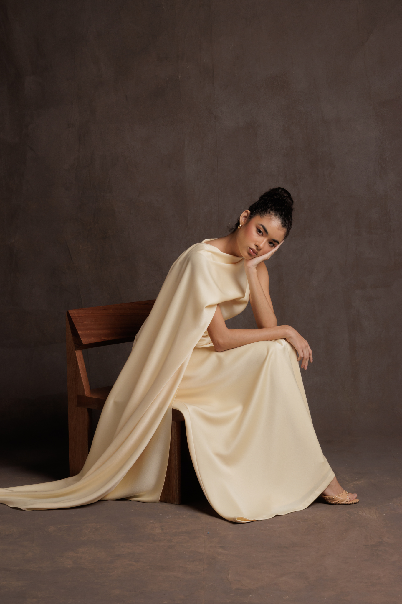 Woman in a flowing beige dress sitting on a wooden chair against a brown background