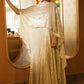 A model stands in a warm-toned interior wearing a shimmering champagne-gold gown with a draped cape-like overlay and a long pleated skirt. Two arched mirrors with patterned glass are behind her, and a wood floor is visible.