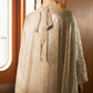 A woman in profile wearing a shimmering champagne-gold textured cape with a tie at the back, standing in a warmly lit interior hallway with arched windows.