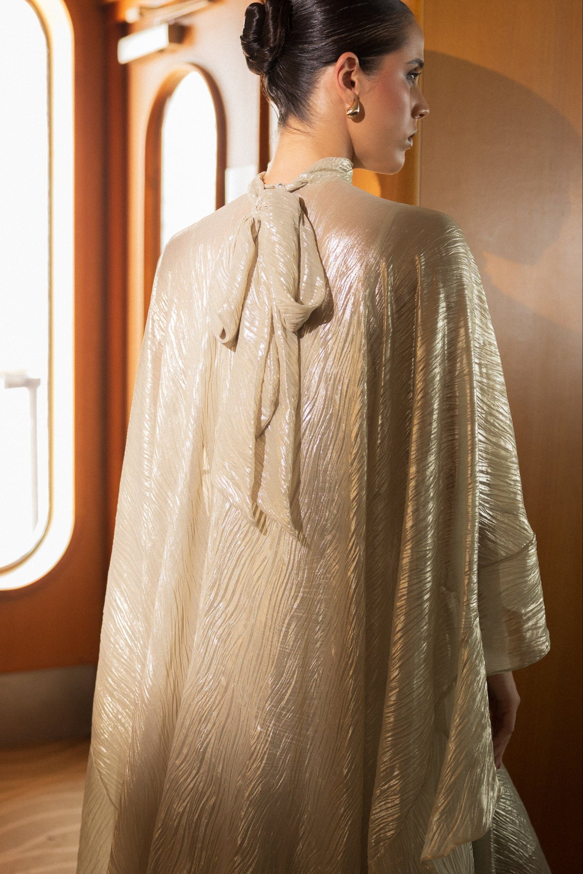 A woman in profile wearing a shimmering champagne-gold textured cape with a tie at the back, standing in a warmly lit interior hallway with arched windows.