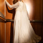 A woman in a long-sleeved cream chiffon gown with intricate lace embroidery on the bodice, posing in a dimly lit elevator with warm wood paneling and a red patterned carpet.