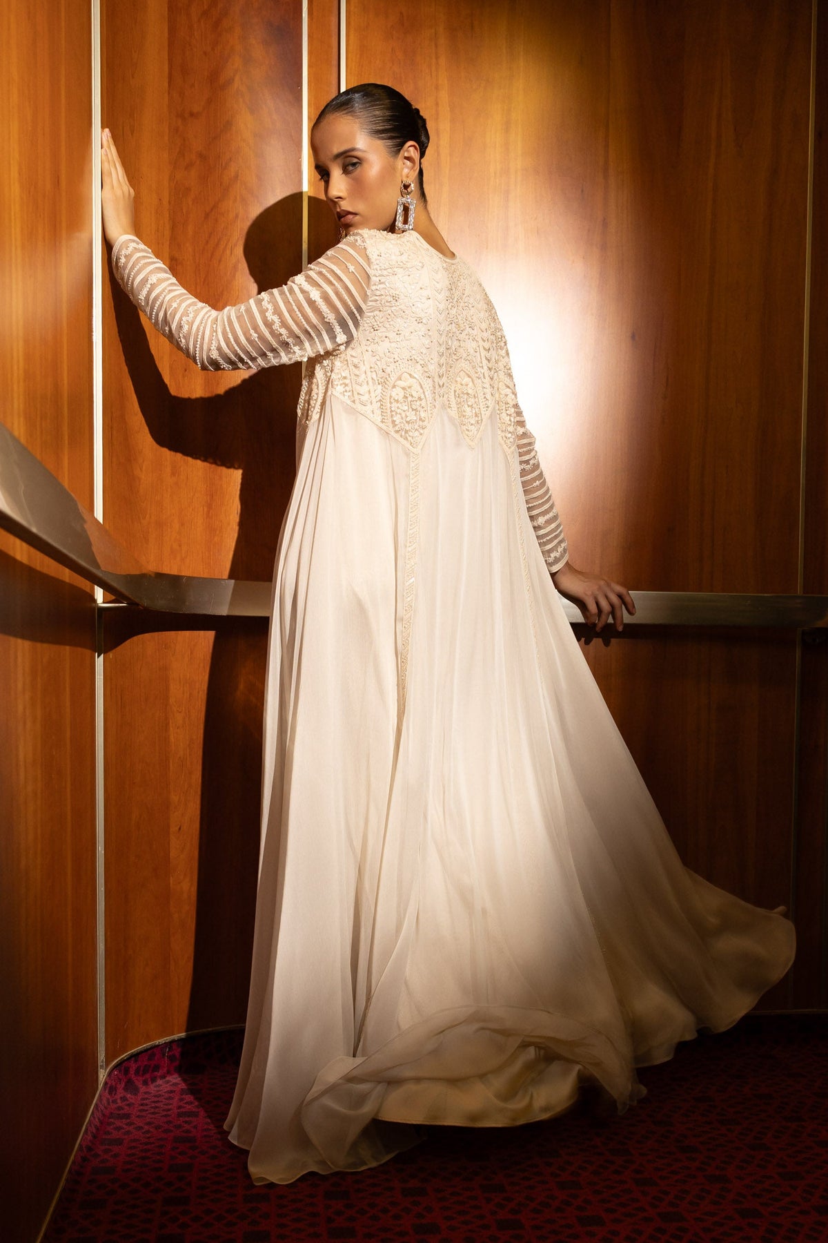 A woman in a long-sleeved cream chiffon gown with intricate lace embroidery on the bodice, posing in a dimly lit elevator with warm wood paneling and a red patterned carpet.