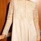 A woman wearing a long-sleeved cream-colored dress with intricate lace and embroidery on the bodice and sleeves, standing in front of a wooden door.