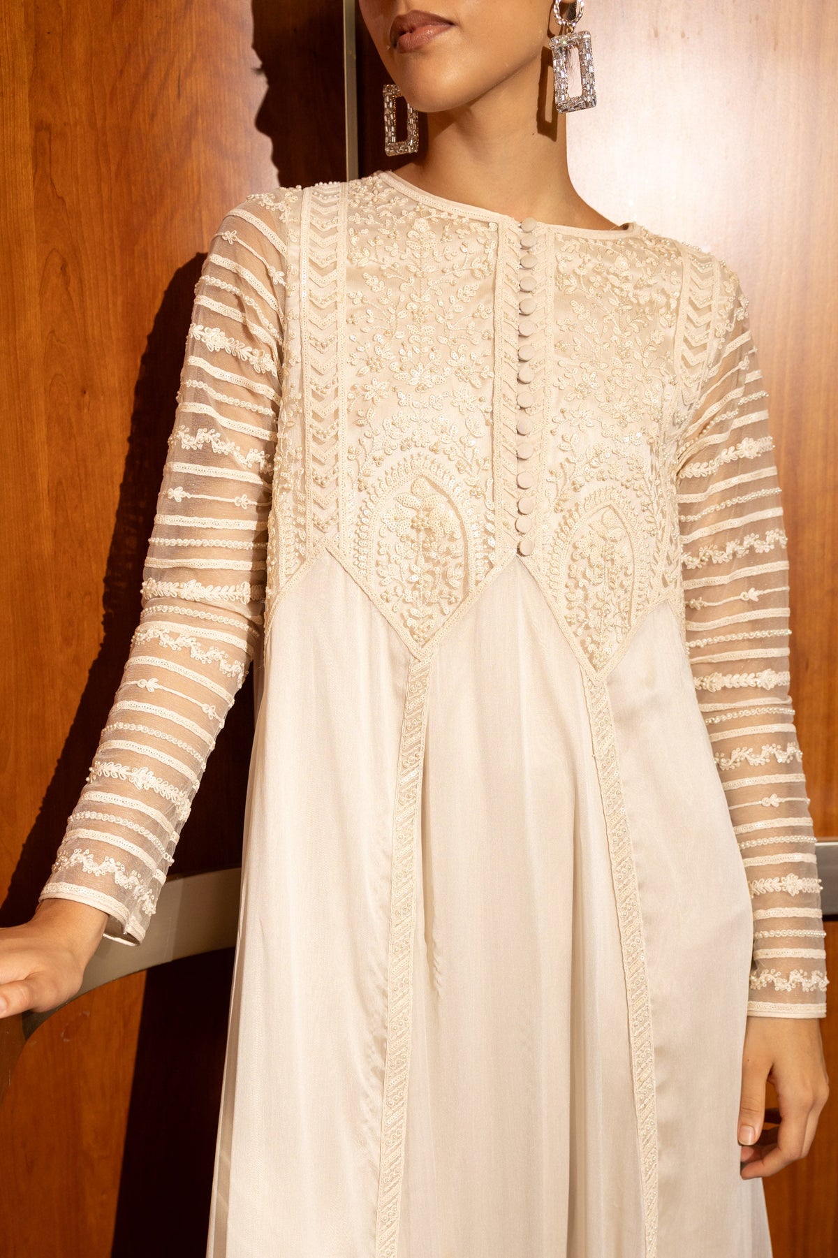 A woman wearing a long-sleeved cream-colored dress with intricate lace and embroidery on the bodice and sleeves, standing in front of a wooden door.