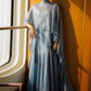 A model stands in a sunlit interior wearing a flowing light blue high-neck maxi dress with draped sleeves and a long train, posed beside a window against a warm orange wall.