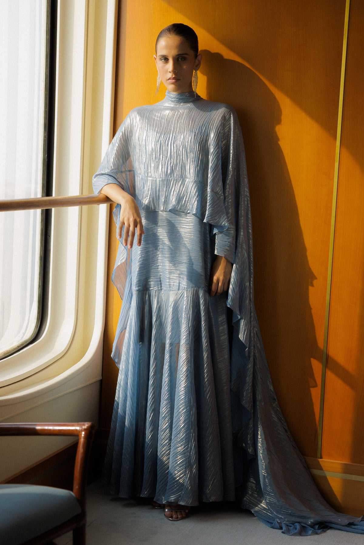 A model stands in a sunlit interior wearing a flowing light blue high-neck maxi dress with draped sleeves and a long train, posed beside a window against a warm orange wall.