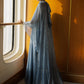 A woman stands by a large window wearing a flowing, floor-length blue gown with sheer draped sleeves, set against a warm wood-paneled wall and floor.