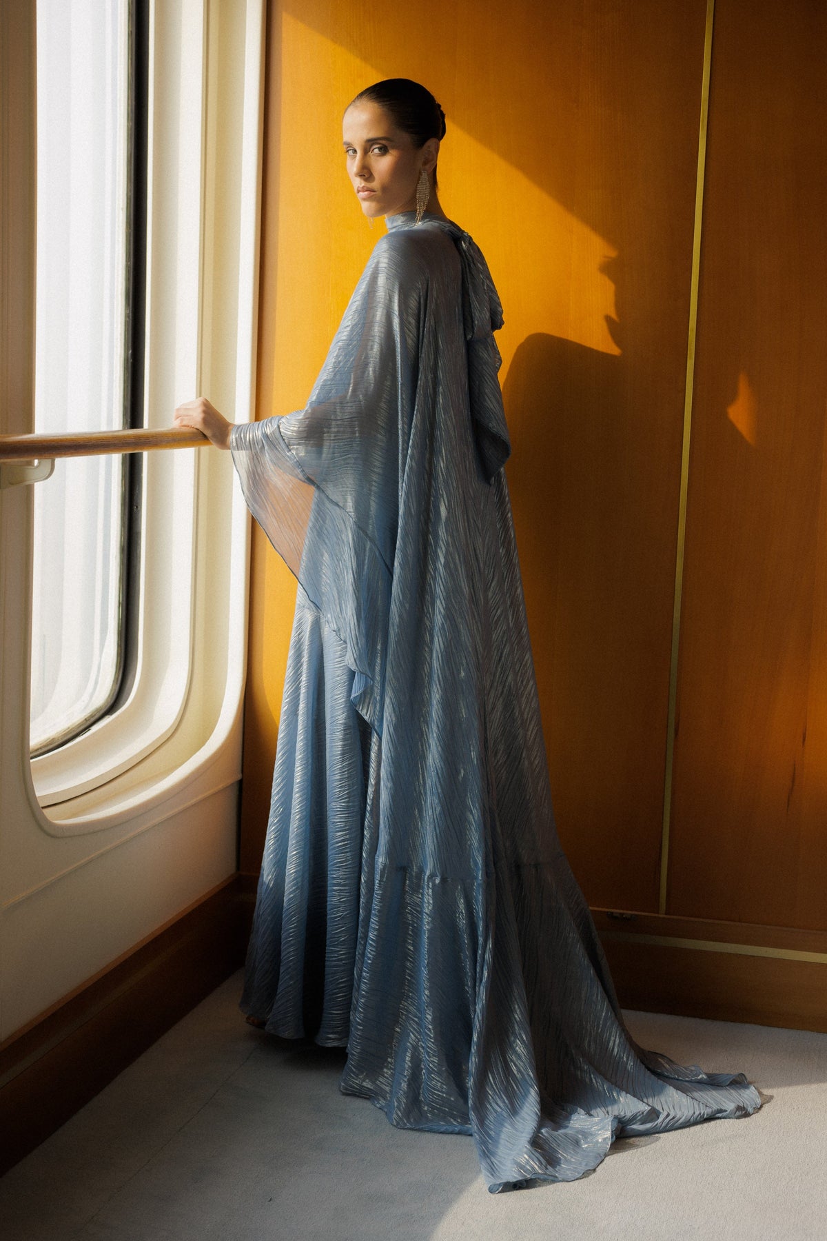 A woman stands by a large window wearing a flowing, floor-length blue gown with sheer draped sleeves, set against a warm wood-paneled wall and floor.