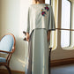 A woman stands indoors near tall arched windows wearing a long flowing dress or robe in a pale sage green color with colorful floral embroidery on the upper left side. The room has warm cream walls, wooden trim, a blue upholstered chair at left, and soft natural light casting shadows on the wall.
