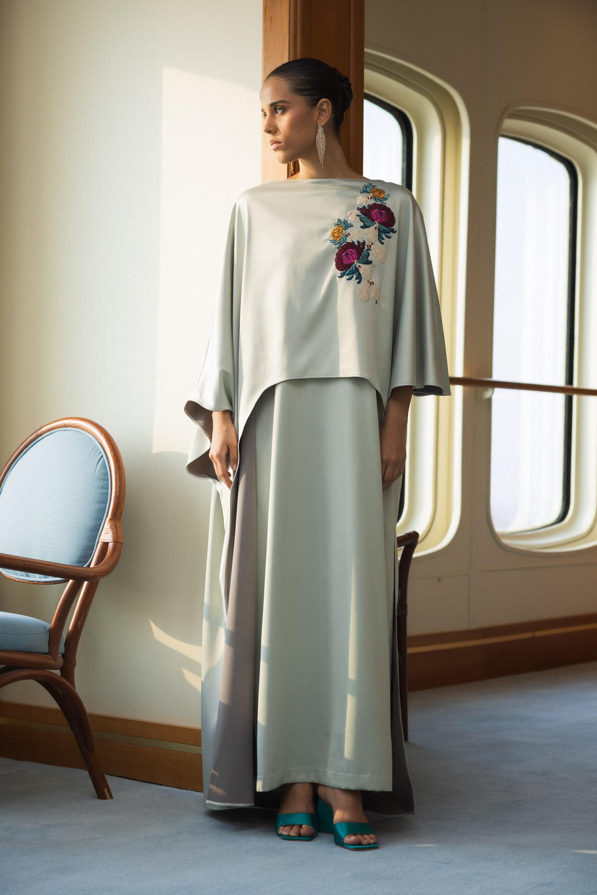 A woman stands indoors near tall arched windows wearing a long flowing dress or robe in a pale sage green color with colorful floral embroidery on the upper left side. The room has warm cream walls, wooden trim, a blue upholstered chair at left, and soft natural light casting shadows on the wall.