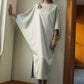 A woman wearing a flowing, light sage green maxi dress with wide draped sleeves and a small floral embroidery on the chest, standing in a room with warm sunlight and a wooden archway.