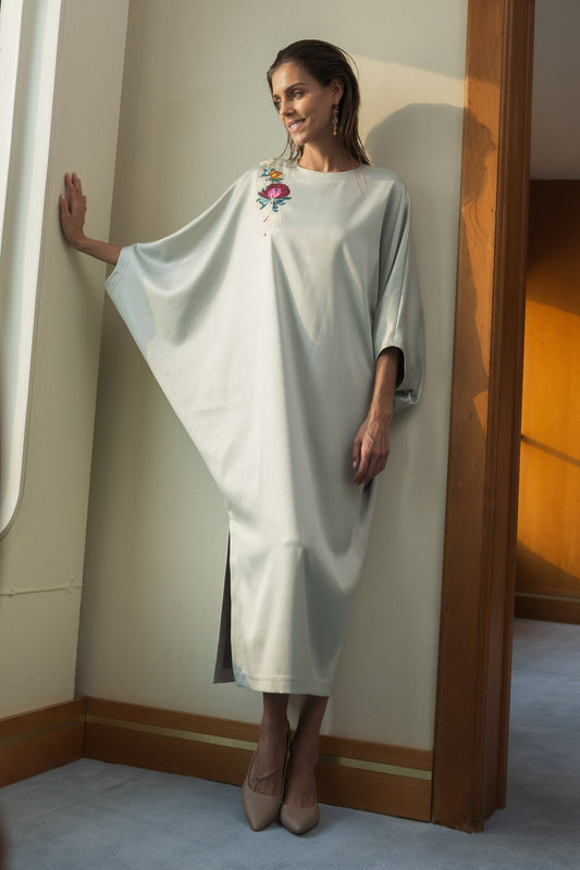 A woman wearing a flowing, light sage green maxi dress with wide draped sleeves and a small floral embroidery on the chest, standing in a room with warm sunlight and a wooden archway.