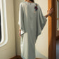 A woman stands in a sunlit interior wearing a flowing, loose-fitting kaftan dress. The dress is a very light sage green with a small floral embroidery on the upper back.