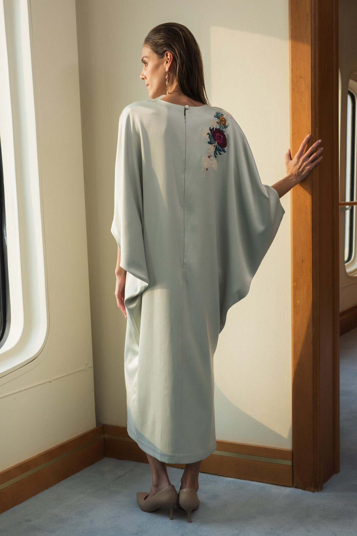 A woman stands in a sunlit interior wearing a flowing, loose-fitting kaftan dress. The dress is a very light sage green with a small floral embroidery on the upper back.