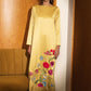 A woman stands in a warm-toned interior wearing a long-sleeved yellow maxi dress with colorful floral embroidery near the hem. A mustard-colored sofa is visible on the left, and the background features wood-paneled walls and a dark blue carpet.