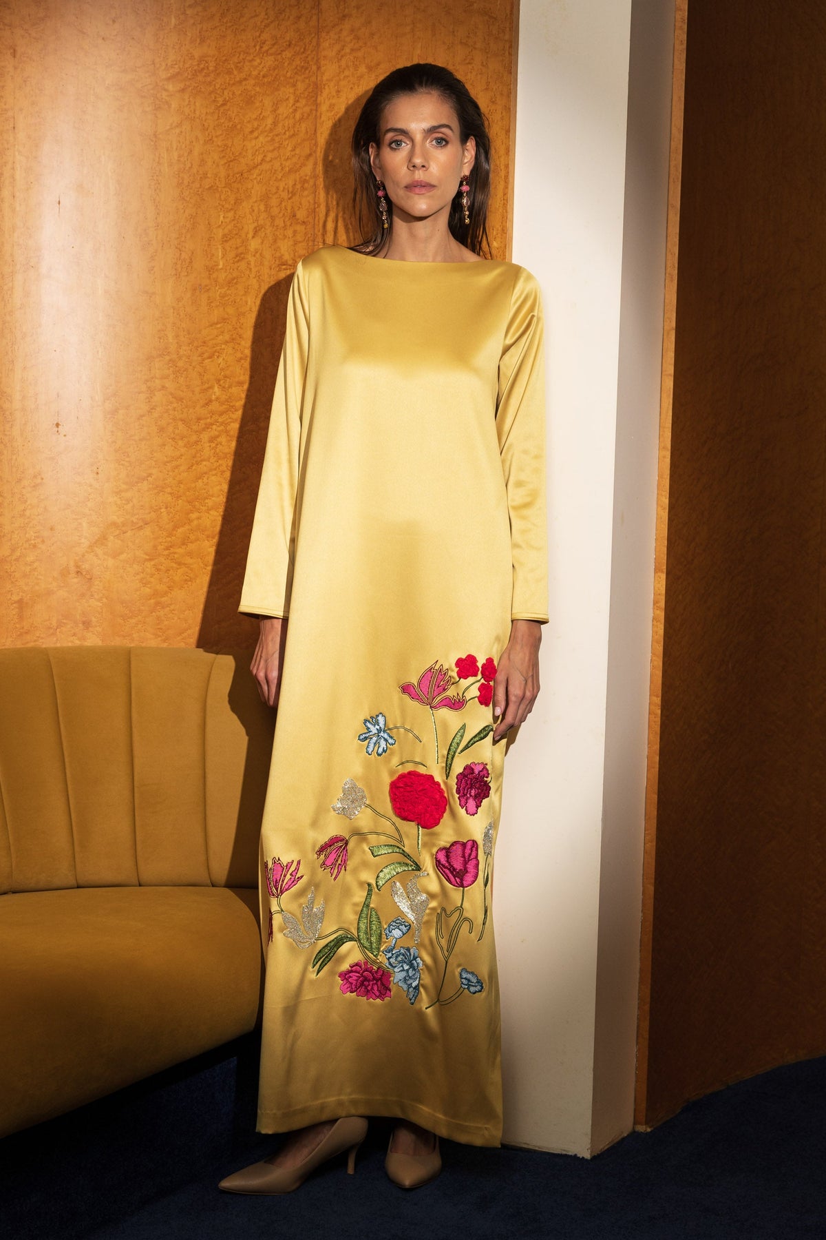 A woman stands in a warm-toned interior wearing a long-sleeved yellow maxi dress with colorful floral embroidery near the hem. A mustard-colored sofa is visible on the left, and the background features wood-paneled walls and a dark blue carpet.