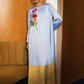 A woman stands indoors wearing a long light blue abaya dress with a gold satin lower skirt and red floral embroidery on the chest. The setting features a warm wood-paneled wall, a mustard-colored sofa, and a dark blue carpet.