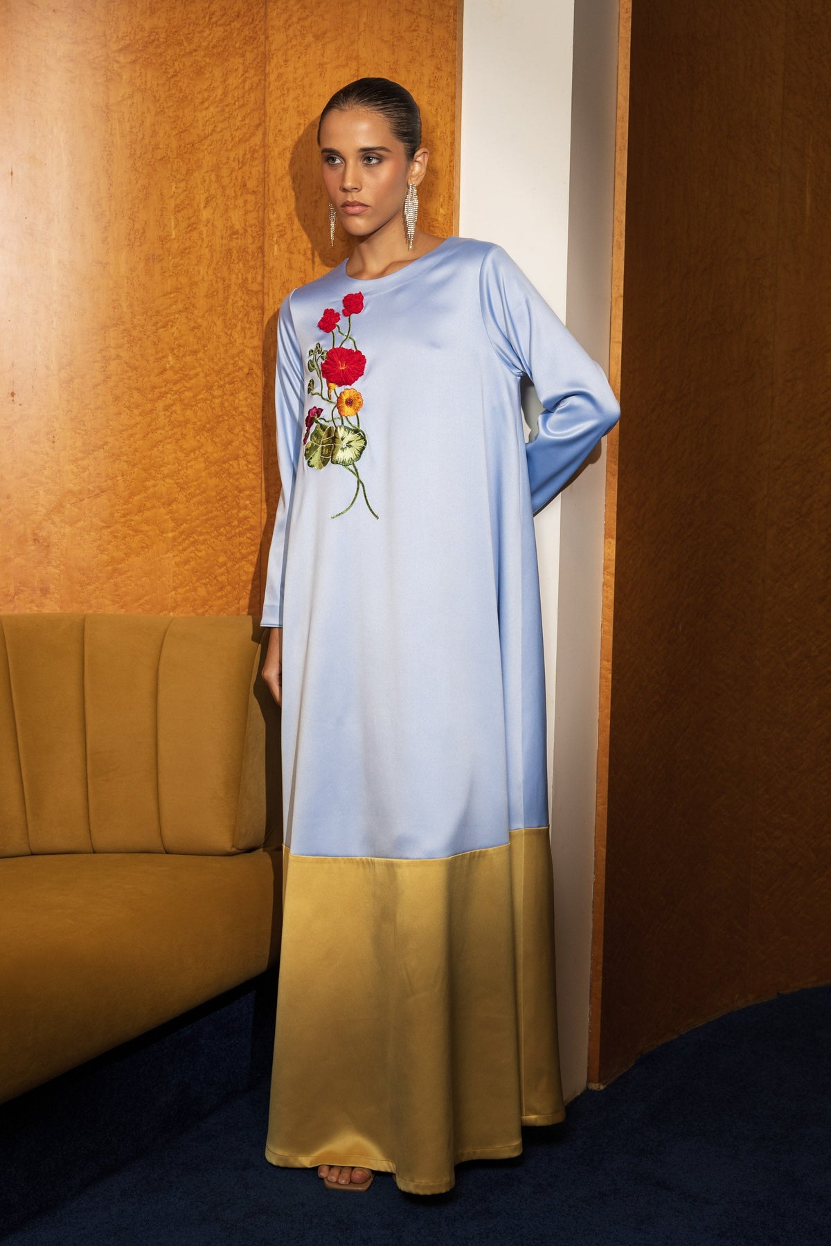 A woman stands indoors wearing a long light blue abaya dress with a gold satin lower skirt and red floral embroidery on the chest. The setting features a warm wood-paneled wall, a mustard-colored sofa, and a dark blue carpet.