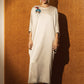 A woman stands against a textured orange-brown wall wearing a loose, knee-length cream dress with a colorful embroidered motif on the upper chest. The scene is lit by direct sunlight casting a strong shadow on the wall, and the floor is dark blue.
