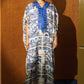 A model stands against a warm brown wood-paneled wall wearing a long, loose caftan dress with a blue and cream architectural pattern, set against a dark blue carpet.