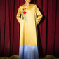 A woman stands in front of a red curtain wearing a long-sleeved dress with a yellow upper half, a light blue lower half, and red floral embroidery on the chest.