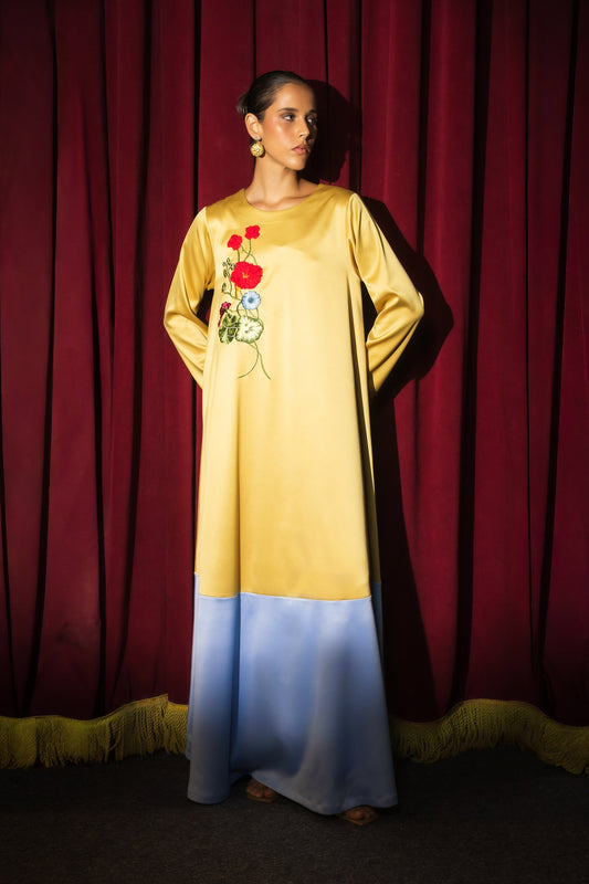 A woman stands in front of a red curtain wearing a long-sleeved dress with a yellow upper half, a light blue lower half, and red floral embroidery on the chest.